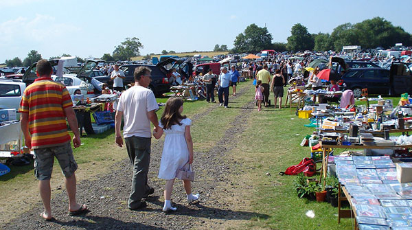 A group of people are walking down a path at a car boot sale. The sun is shining and there are lots of stalls with different items on for sale.