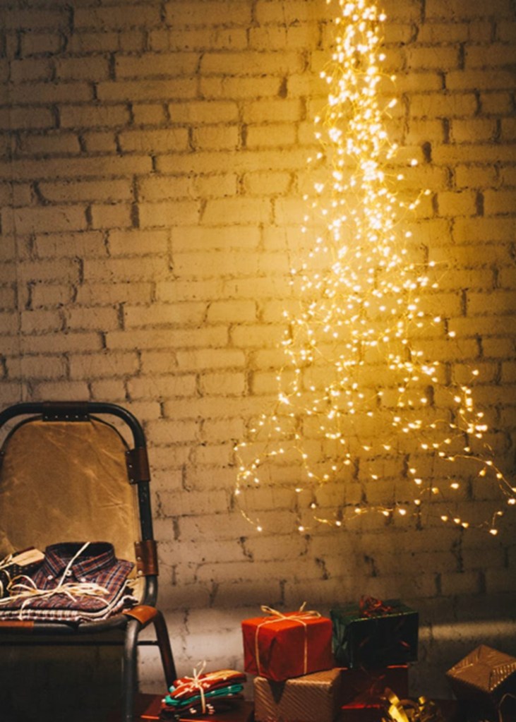 A white brick wall decorated with golden fairy lights, strung up in the shape of a Christmas tree. At the base is a chair and a small pile of presents.