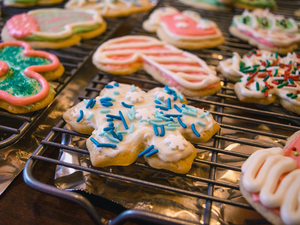 Brightly decorated festive biscuits, including a snowflake biscuit with blue sprinkles and a candy-cane biscuit with pink and white iced stripes, arranged neatly on a cooling rack. 