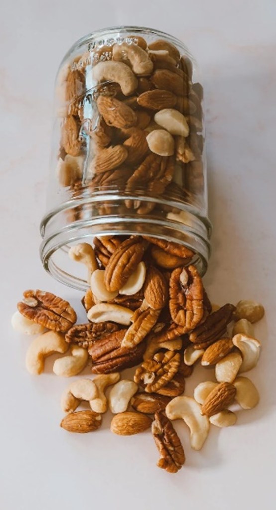 A jar containing nuts on a white background that has been tipped over, spilling its contents. The nuts include pecans, cashews, and almonds.